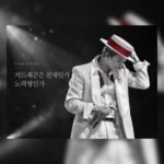 흑백 무대 조명 아래 붉은 띠가 있는 모자를 잡고 고개를 숙인 지드래곤 공연 사진
